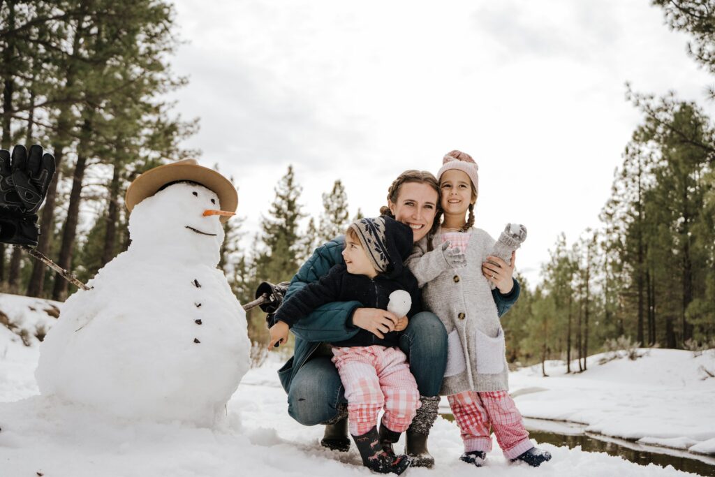 adulte et deux enfants souriants près d'un bonhomme de neige en forêt hivernale