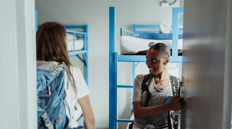 Two women with backpacks walking into a bright hostel dormitory with bunk beds, ready for adventure.